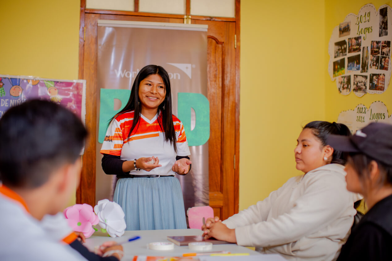 A girl stands at the end of a table, talking to a group of people sitting at the table.